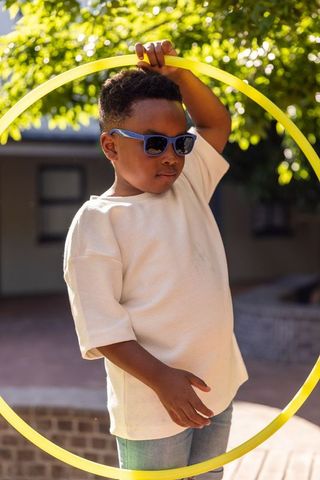 African american boy wearing sunglasses playing with yellow hula hoop in sunlit courtyard