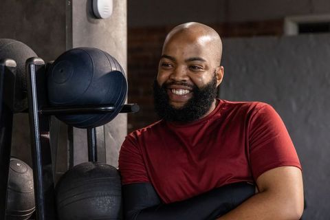 Smiling african american man leaning by medicine ball rack in gym wearing red top bearded