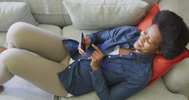 This image depicts an African American woman relaxing on a sofa while using her smartphone. The setting is casual and comfortable, making it ideal for use in articles or advertisements related to home life, comfort, technology use, social media interaction, or leisure time. It is also suitable for lifestyle blogs, digital communication campaigns, or promotional material for mobile devices and internet services.