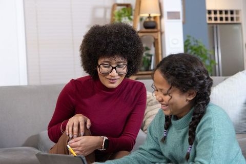 African american mother guiding daughter using tablet, smiling in cozy living room