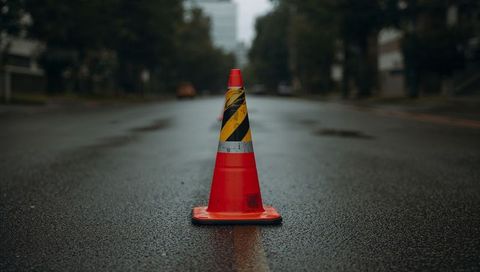 Orange traffic cone standing on wet urban road with reflective band and hazard tape