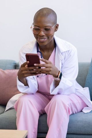 African woman clinician scrolling smartphone in pink scrubs and white coat, smiling