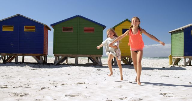 Happy caucasian girls in swimsuits running and smiling near wooden houses on sunny beach. Childhood, friendship, free time, summer, travel, vacations and lifestyle, unaltered.