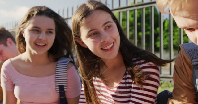 Teenage friends are enjoying an outdoor conversation by a metal fence on a sunny day. They are dressed in casual attire and appear happy, sharing a joyful moment. This could be perfect for illustrating teenage friendship, youth lifestyle, or summer activities.