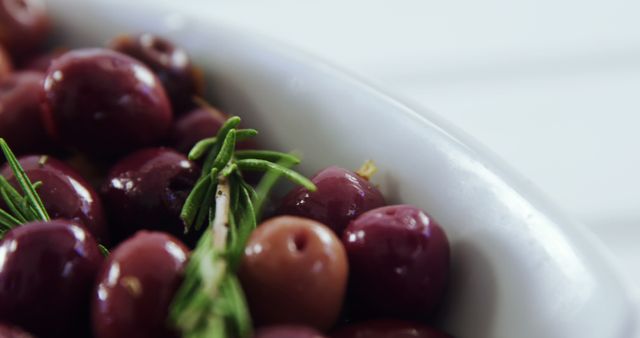 Vibrant close-up of juicy red olives with a sprig of fresh rosemary in a white bowl. Perfect for illustrating topics related to healthy eating, gourmet Mediterranean cuisine, vegan and vegetarian diets, and organic produce. Ideal for use in recipe blogs, health food publications, diet-related articles, and culinary websites.