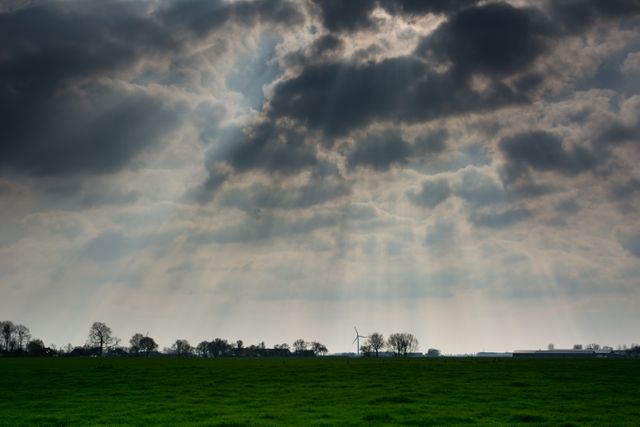 This scene captures sunbeams breaking through dark clouds and illuminating a lush green field. Trees dot the horizon line, and a wind turbine stands to the right, blending naturally into the landscape. Ideal for use in projects emphasizing the beauty of nature, renewable energy, tranquility, or dramatic weather. Perfect for backgrounds, environmental themes, and inspirational contexts.