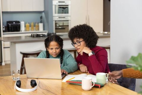 Caregiver tutoring school-age girl on laptop at kitchen table, homeschooling support