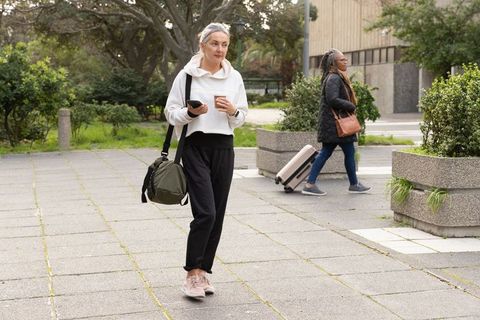 Mature woman walking with coffee and duffel while traveler rolling suitcase in urban plaza
