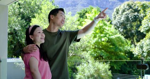 Asian couple standing on balcony pointing at mountain ridge with lush green terrace view