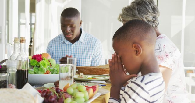 Family prays before enjoying meal outdoors. Two adults and a child bow heads in prayer around a table with fresh food. Suitable for themes of family, faith, and eating together. Useful for advertisements, articles, blogs about family values, gratitude, and religious practices.
