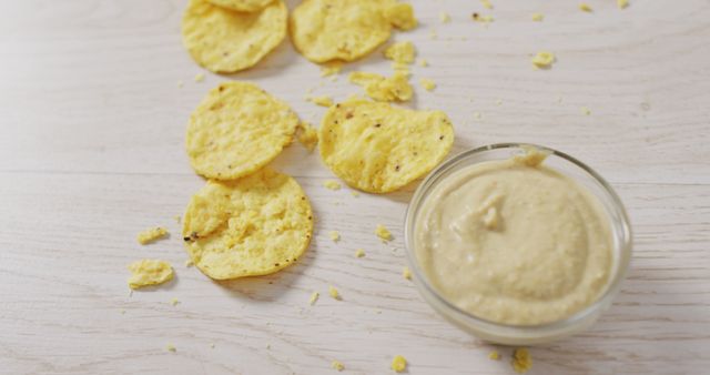 Tortilla chips spread on wooden surface with a bowl of creamy hummus dip. Ideal for ads or articles about snacks, appetizers, parties, or casual dining. Perfect for social media posts, blogs, or recipes focused on healthy snacking options.