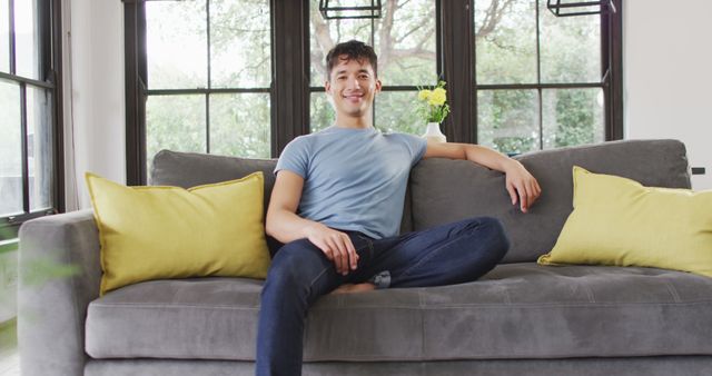 A young man is casually sitting on a stylish grey sofa in a modern and bright living room with large windows. He is wearing a light blue t-shirt and dark jeans, smiling as he looks at the camera. This image is perfect for themes related to home comfort, relaxation, modern lifestyles, and youthful living spaces.