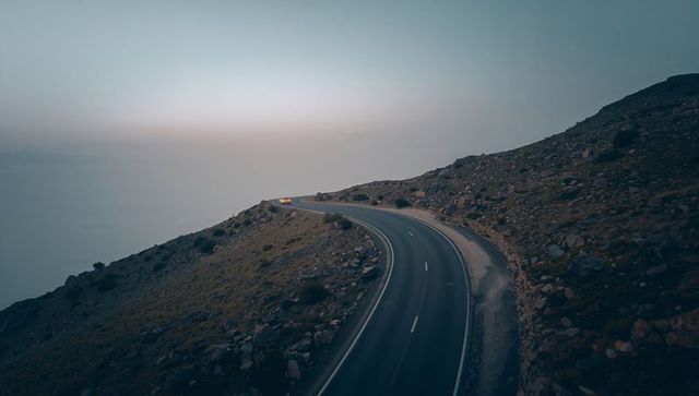 Winding coastal road at dusk with headlights guiding through misty rocky hillside