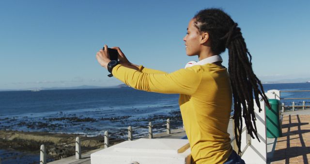 This scene captures a person immersed in photography by the sea, highlighting the joy of outdoor travel and digital engagement. Perfect for representing leisure activities, travel inspirations, digital nomadism, and mindfulness by natural surroundings. Use in travel blogs, lifestyle magazines, or any content focused on the serene experience of nature and innovation.