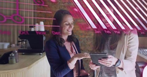 Businesswomen collaborating over tablet in cafe with espresso bar and pink data overlay
