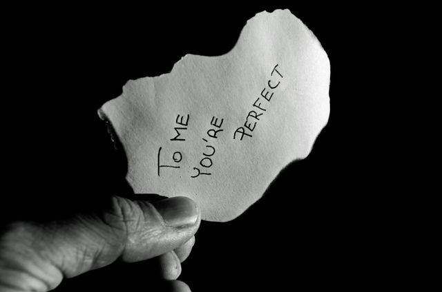 Hand holding a piece of ripped paper with a handwritten message saying 'To me you're perfect', against a black background. Ideal for Valentine's Day promotions, love-themed designs, relationship blogs, greeting cards, and minimalist artworks that emphasize the power of words and simple gestures.
