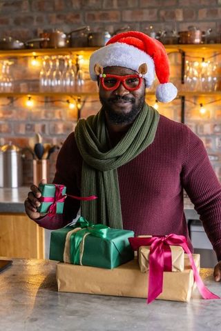 African american man wearing santa hat holding gifts in cozy kitchen with string lights