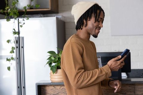 African American man checking phone in kitchen with plant, wearing beanie and pullover