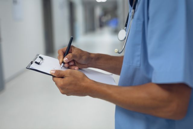 Male surgeon in blue scrubs writing on a clipboard in a hospital corridor. Ideal for use in healthcare, medical, and hospital-related content. Can be used in articles, blogs, and promotional materials about healthcare professionals, medical documentation, and patient care.