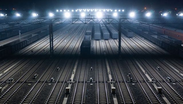Floodlit nighttime freight yard with parallel tracks and parked wagons casting long shadows