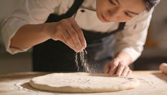 Asian chef sprinkling flour on pizza dough closeup professional baker preparing handcrafted crust