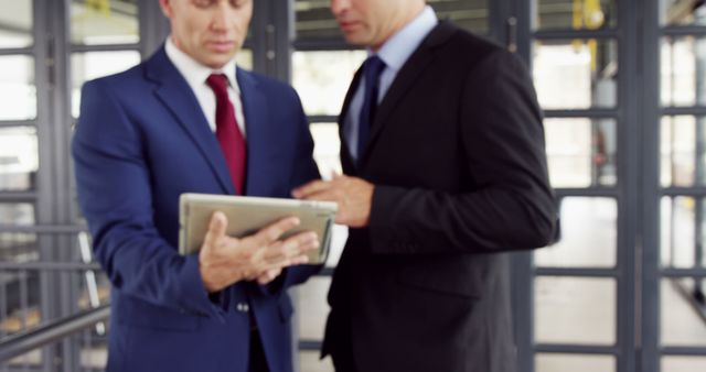 Two businessmen in suits are engaged in a discussion over a digital tablet, with copy space. Their professional attire and focused interaction suggest a corporate setting where important information is being shared or reviewed.
