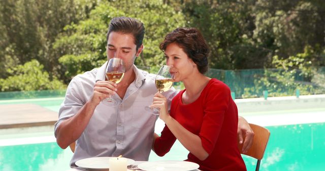 A Caucasian couple enjoys a romantic outdoor meal by a pool, toasting with glasses of white wine. Their elegant attire and the serene setting suggest a special occasion or a luxurious retreat.