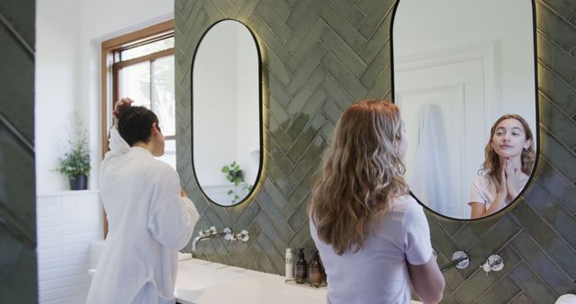 Women are seen engaging in their morning routines in a stylish, modern bathroom. The space features green tiled walls, large oval mirrors, and a window allowing natural light. Perfect for illustrating morning rituals, self-care, skincare routines, and modern interior design inspirations.
