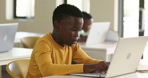 Young African boy typing on laptop while studying in bright classroom, focused on learning
