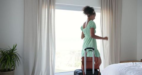Young woman with suitcase by window in bright bedroom interior