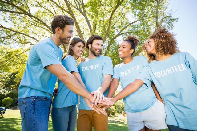 Group of diverse volunteers in blue shirts, stacking hands in unity. Ideal for community service promotions, charitable organization marketing, teamwork building activities, and diversity inclusion initiatives.