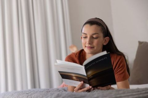 Young woman reading paperback on bed cozy bedroom relaxation with soft natural light
