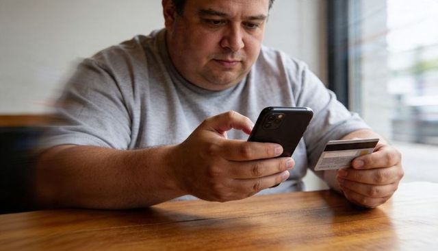 Man using smartphone and credit card for mobile payment at cafe table by window