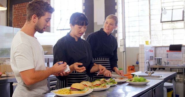 Chef instructing apprentices on food preparation in a modern industrial kitchen. Chef pointing at dishes while apprentices listen attentively, showcasing the culinary learning process in a professional environment. Ideal for use in articles about culinary education, team training, restaurant operations, or apprenticeship programs.