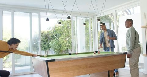 Diverse young men playing pool in bright modern game room with floor-to-ceiling glass