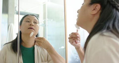 Mature african american woman using rose quartz facial roller at bathroom mirror during self-care ro