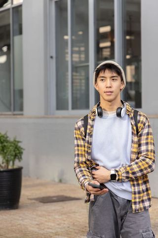 Teen asian student standing campus courtyard checking phone with headphones and backpack