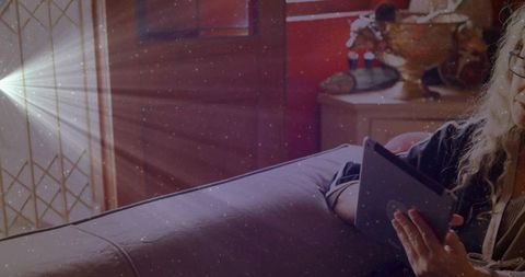 Mature woman reading tablet on sunlit sofa by patterned glass door, cozy home relaxation