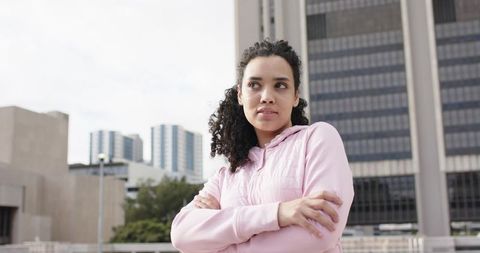 Young woman standing on urban balcony wearing pink hoodie looking thoughtful and confident
