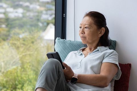 Senior asian woman gazing out window holding smartphone and wearing smartwatch