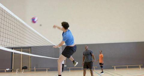 Male athlete jumping and spiking volleyball during indoor gym match with teammates watching