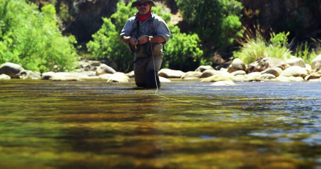 Man fly fishing in a shallow river on a sunny day. He is standing in the water, calmly engaging in the activity while surrounded by nature, including trees and rocks. Ideal for depicting recreational outdoor activities, leisure, and connection with nature, this can be used by websites, blogs about fishing, marketing for fishing gear, or promoting outdoor hobbies and lifestyles.