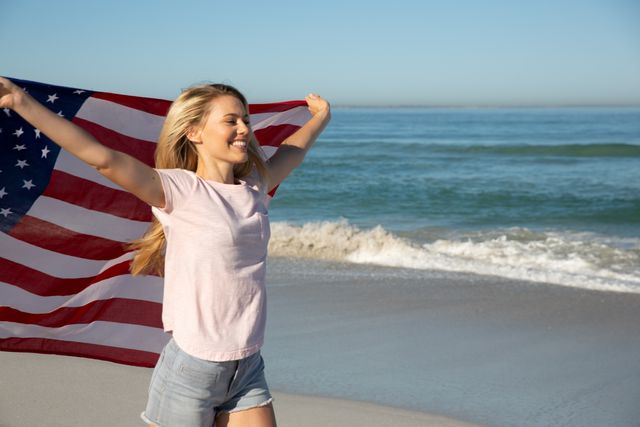 Caucasian woman enjoying time at the beach on a sunny day, smiling and running with an American flag, with sea in the background. Holiday relaxing summer wellbeing.