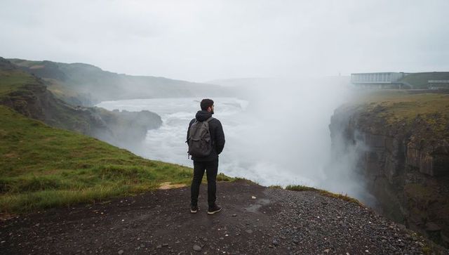 Solo hiker standing at misty waterfall cliff overlooking rugged river gorge