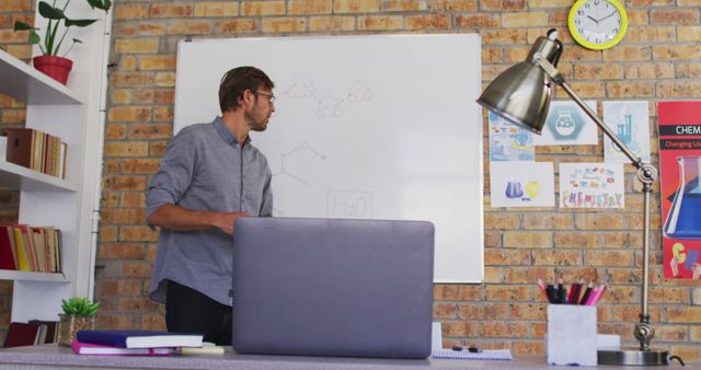 Male teacher presenting chemistry lessons on a whiteboard in a classroom with a brick wall. Ideal for use in educational articles, science-related advertisements, academic materials, and teacher resources.
