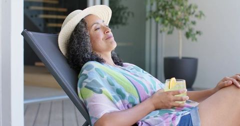 Woman relaxing on patio lounge chair wearing straw hat, sipping lemon drink, sunlit