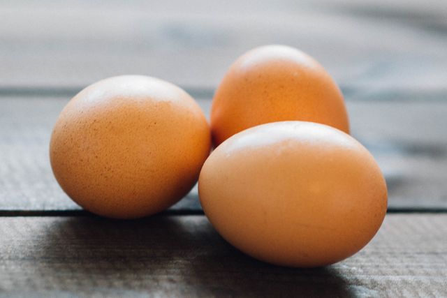 Three brown eggs resting on a wooden surface with soft light falling on them. Warm tones and rustic setting give this image a natural and organic feel, making it ideal for food blogs, farm fresh product promotions, recipes, or nutritional content.
