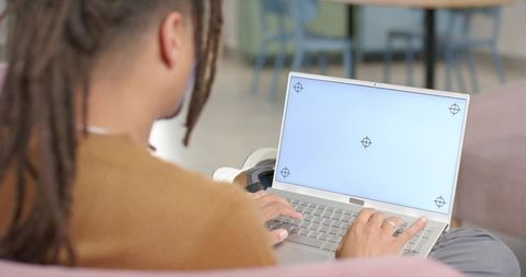 Man with dreadlocks typing on laptop on pink sofa in minimalist home lounge remote work