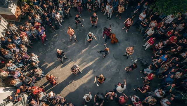 Aerial street performance with buskers, dancers, upright bass and crowded sunlit urban courtyard