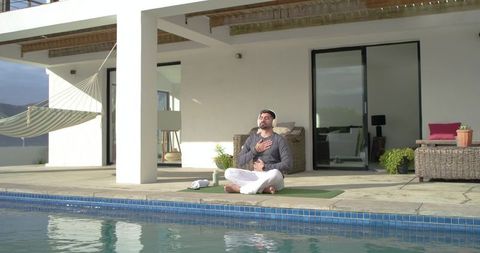 Indian man meditating by pool on modern terrace with yoga mat and headphones